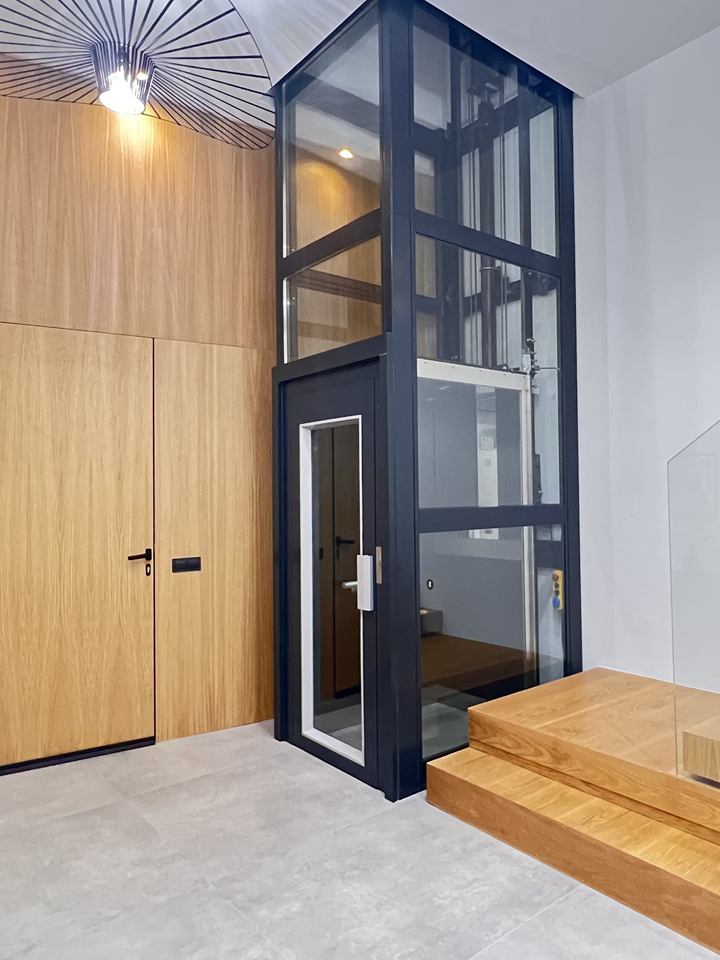 Home lift with a hinged swing door in a black-framed glass shaft beside a floating staircase